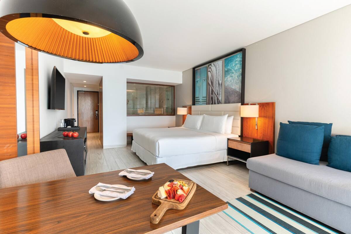A modern hotel room with a bed, a dining table with plates and cutlery, a sofa, a TV mounted on the wall, and a painting above the bed.