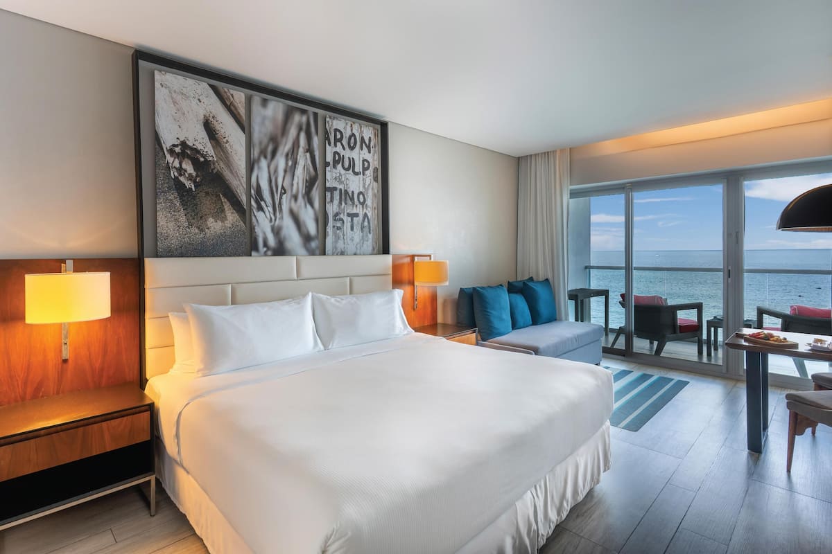 A hotel room with a large bed, a desk with a chair, a sofa, and a large window with a view of the sea.