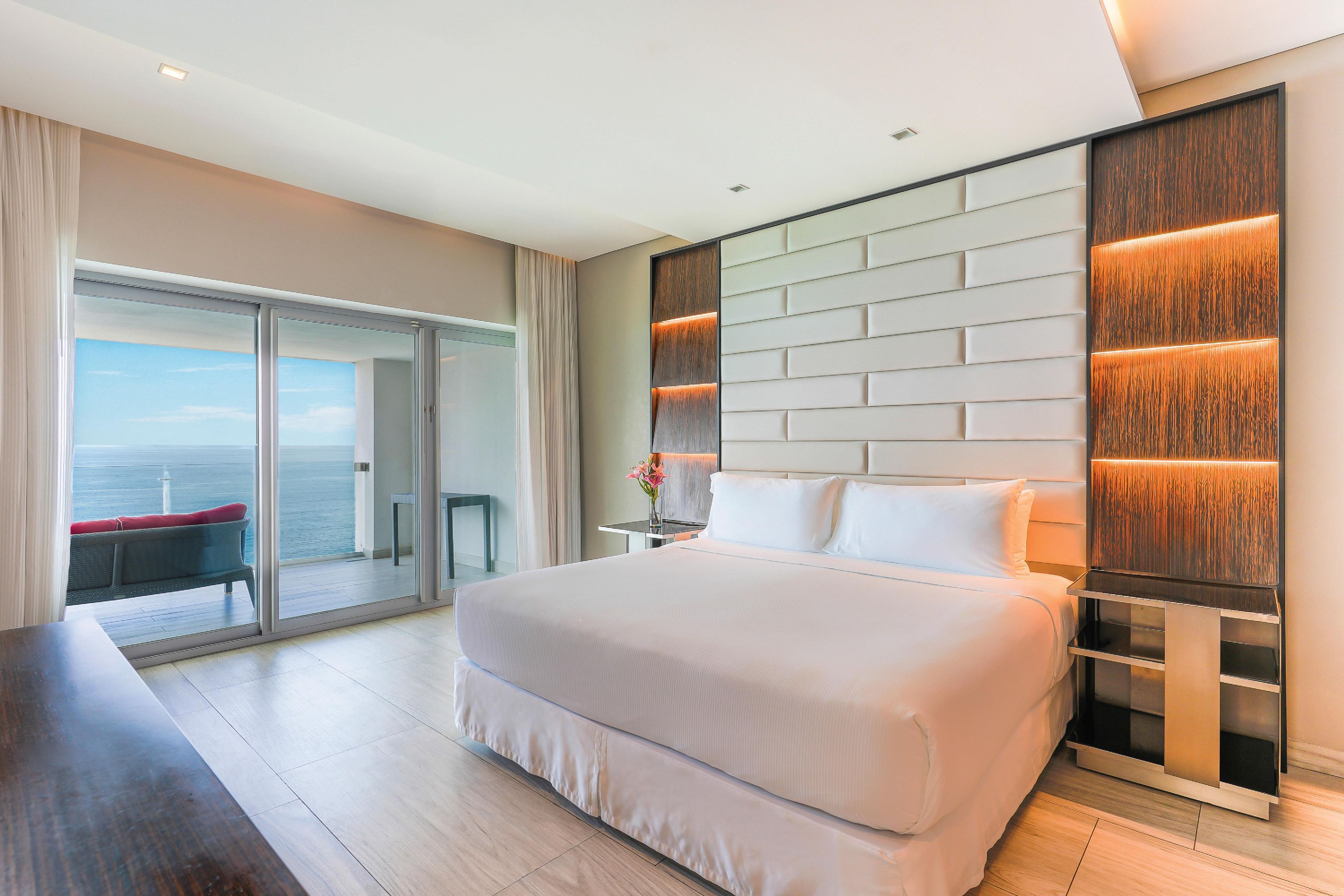 Een moderne hotelkamer met een groot bed, een glazen deur naar een balkon, en een houten accentmuur met ingebouwde verlichting.