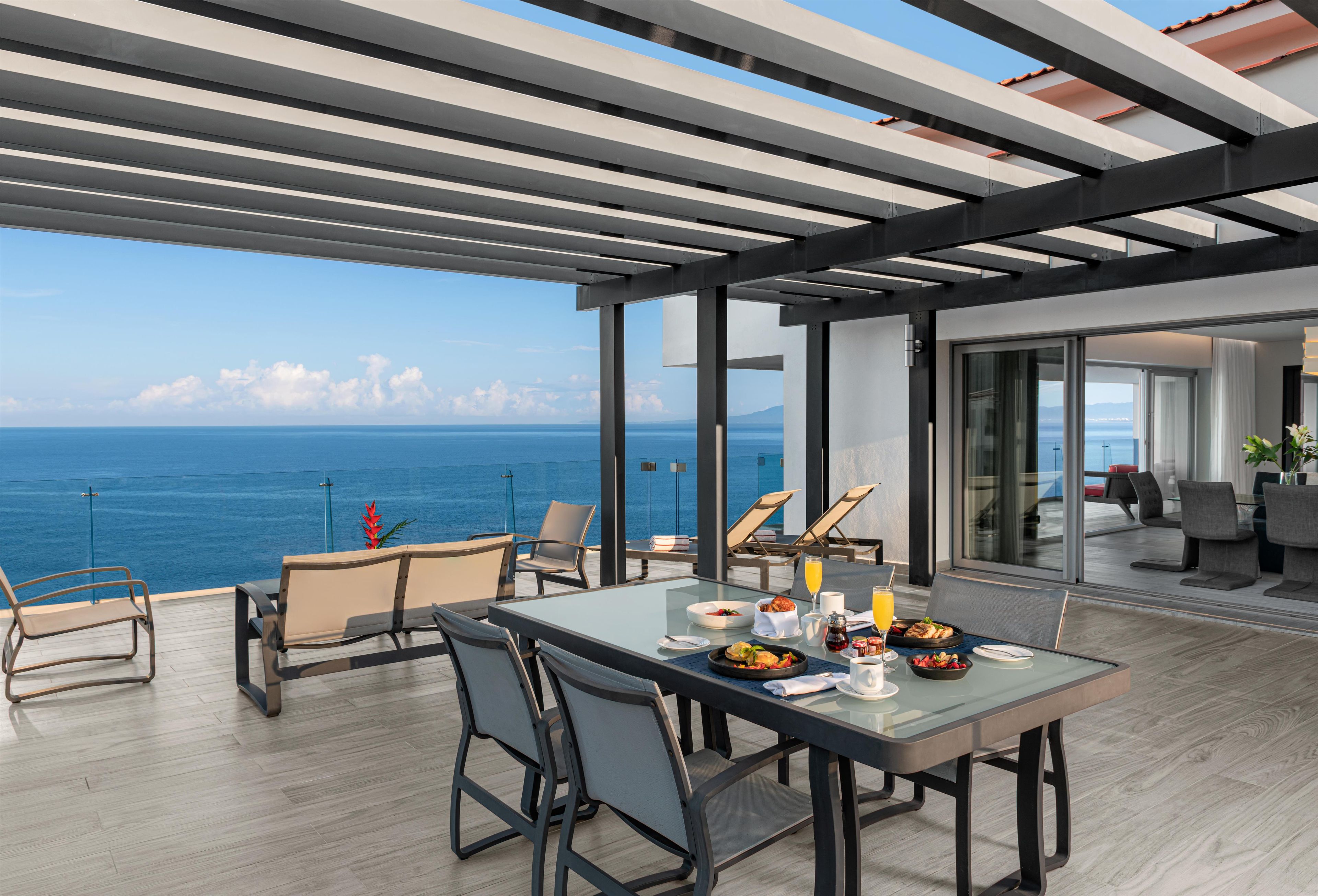 Un área de comedor al aire libre y moderna con vista al mar, que incluye una mesa dispuesta con comida y bebidas, rodeada de sillas.