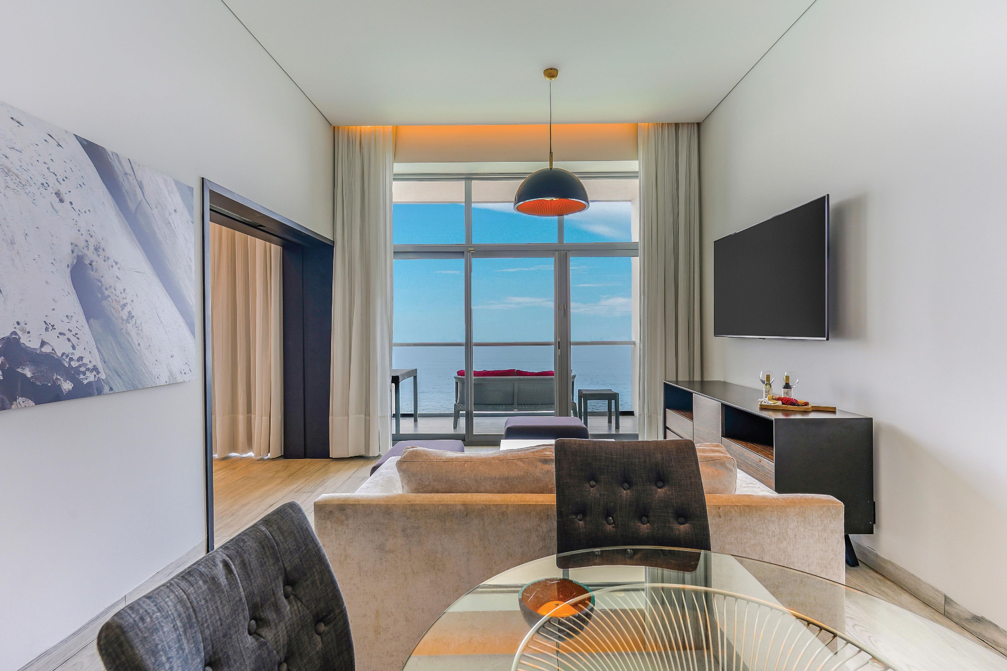 Habitación de hotel moderna con una mesa de vidrio, un sofá, un televisor y un ventanal con vistas al mar.