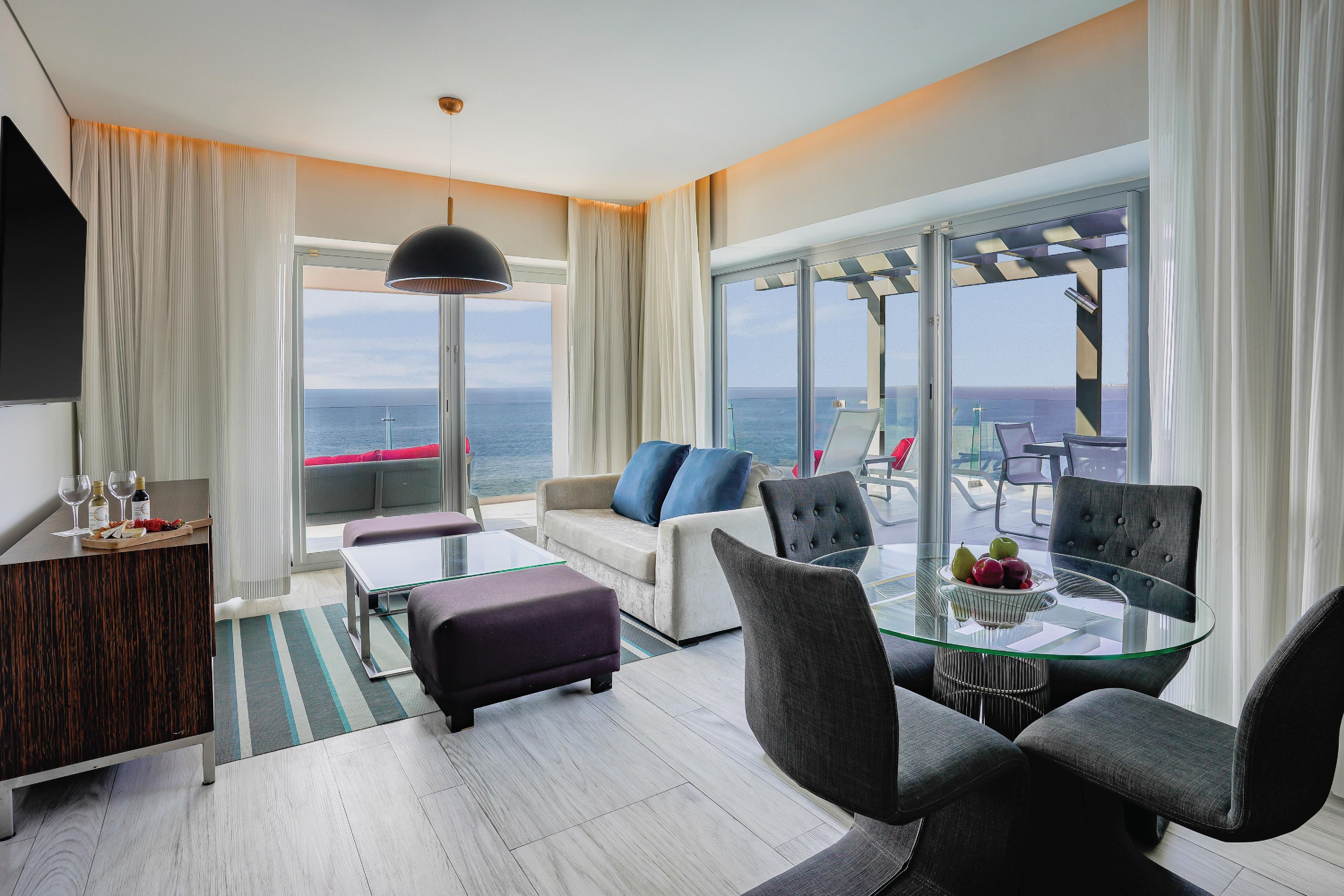 Une chambre d’hôtel moderne avec une table en verre, des sièges confortables, un canapé, une télévision et une vue sur la mer.
