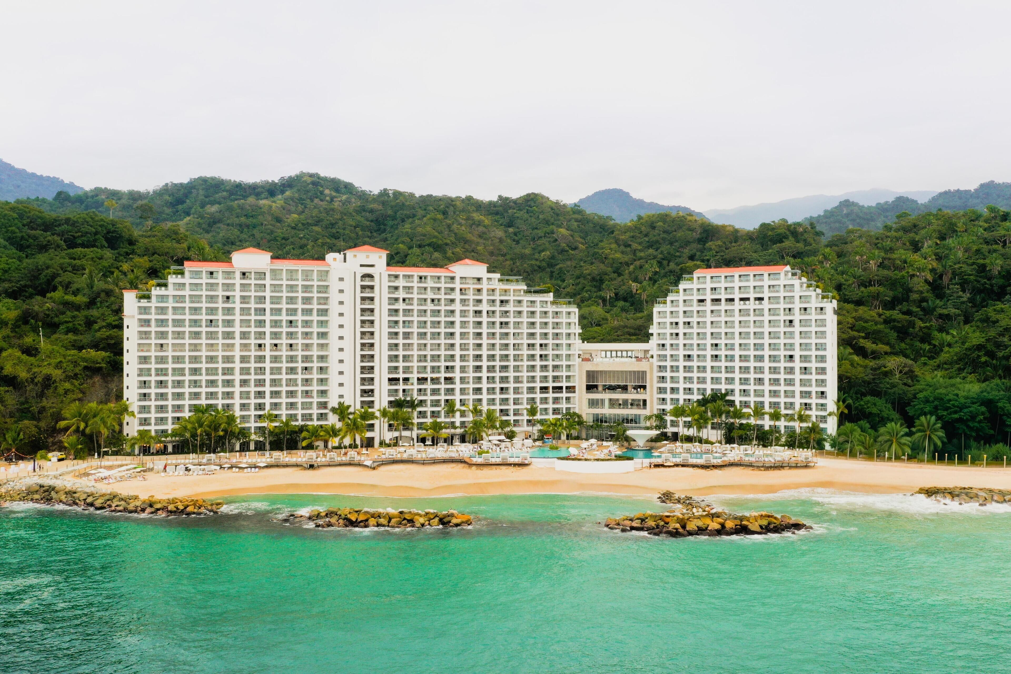 A large hotel with multiple floors, a beach, and a mountainous backdrop.