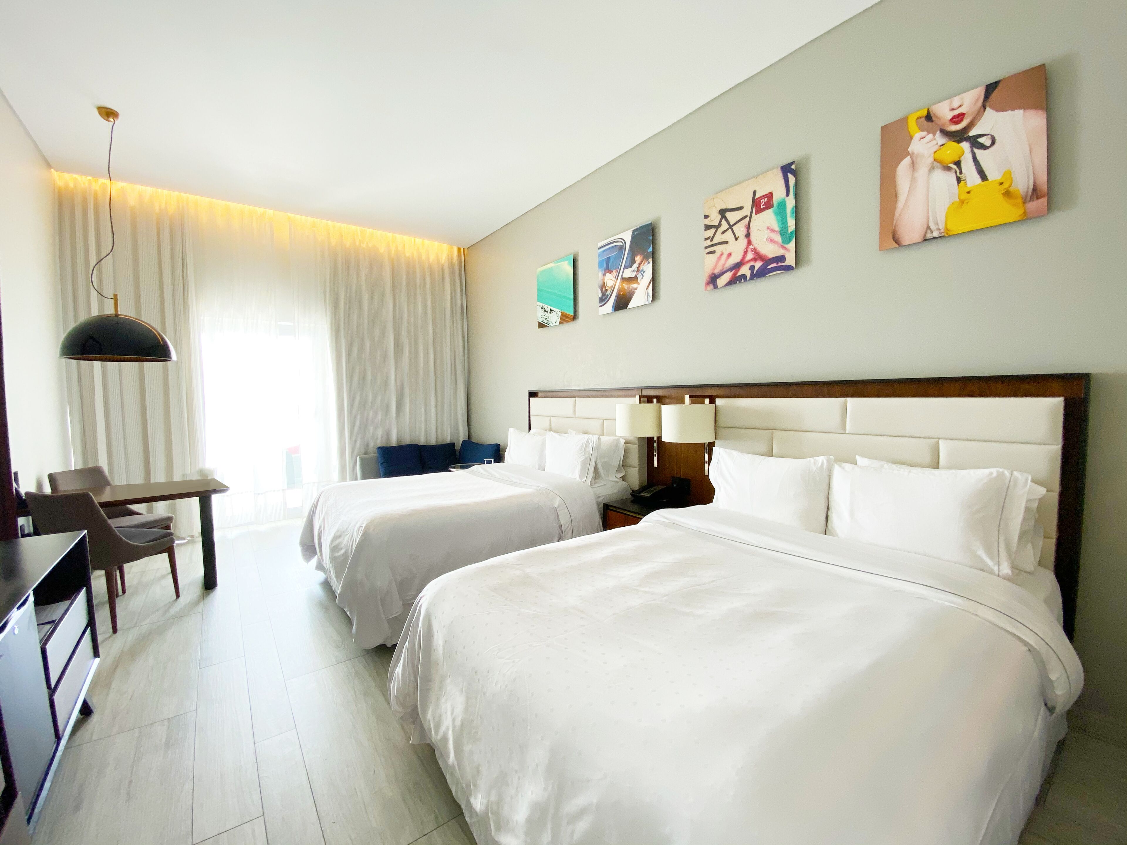 A hotel room with two beds, a desk, a chair, a lamp, and artwork on the wall.