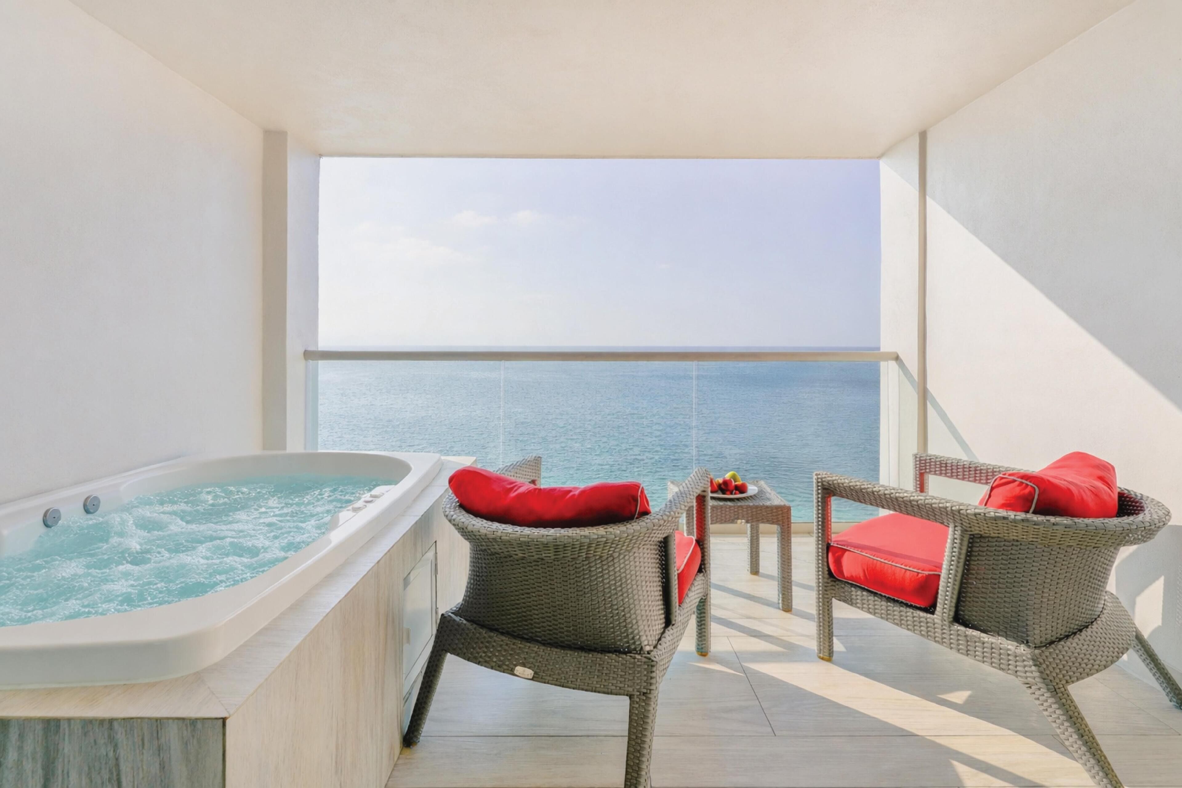 A balcony with a hot tub, wicker chairs with red cushions, and a view of the sea.