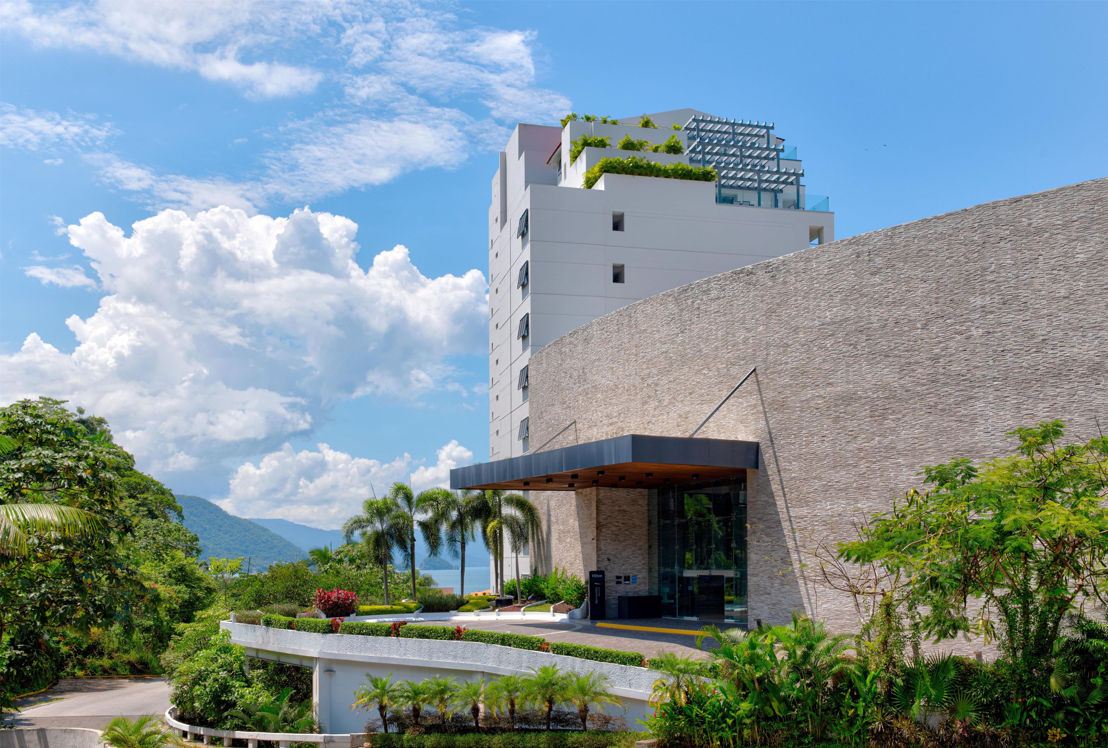 Un hotel moderno con fachada de piedra, techo plano con vegetación y vista al mar.