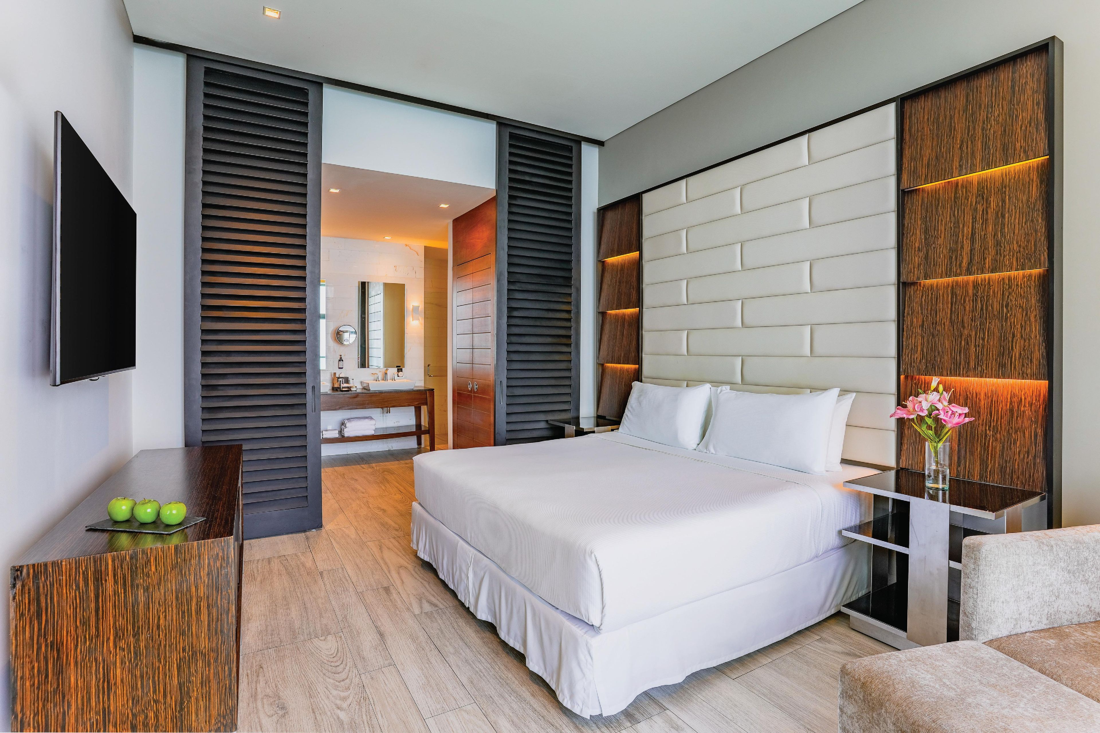 A modern hotel room with a large bed, a flat-screen TV, a wooden desk, and a bathroom visible through an open door.