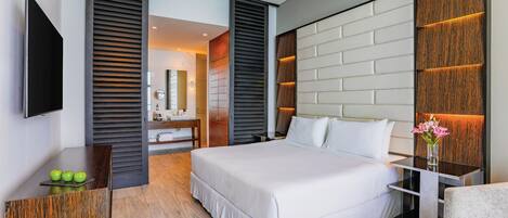 A modern hotel room with a large bed, a flat-screen TV, a wooden desk, and a bathroom visible through an open door.