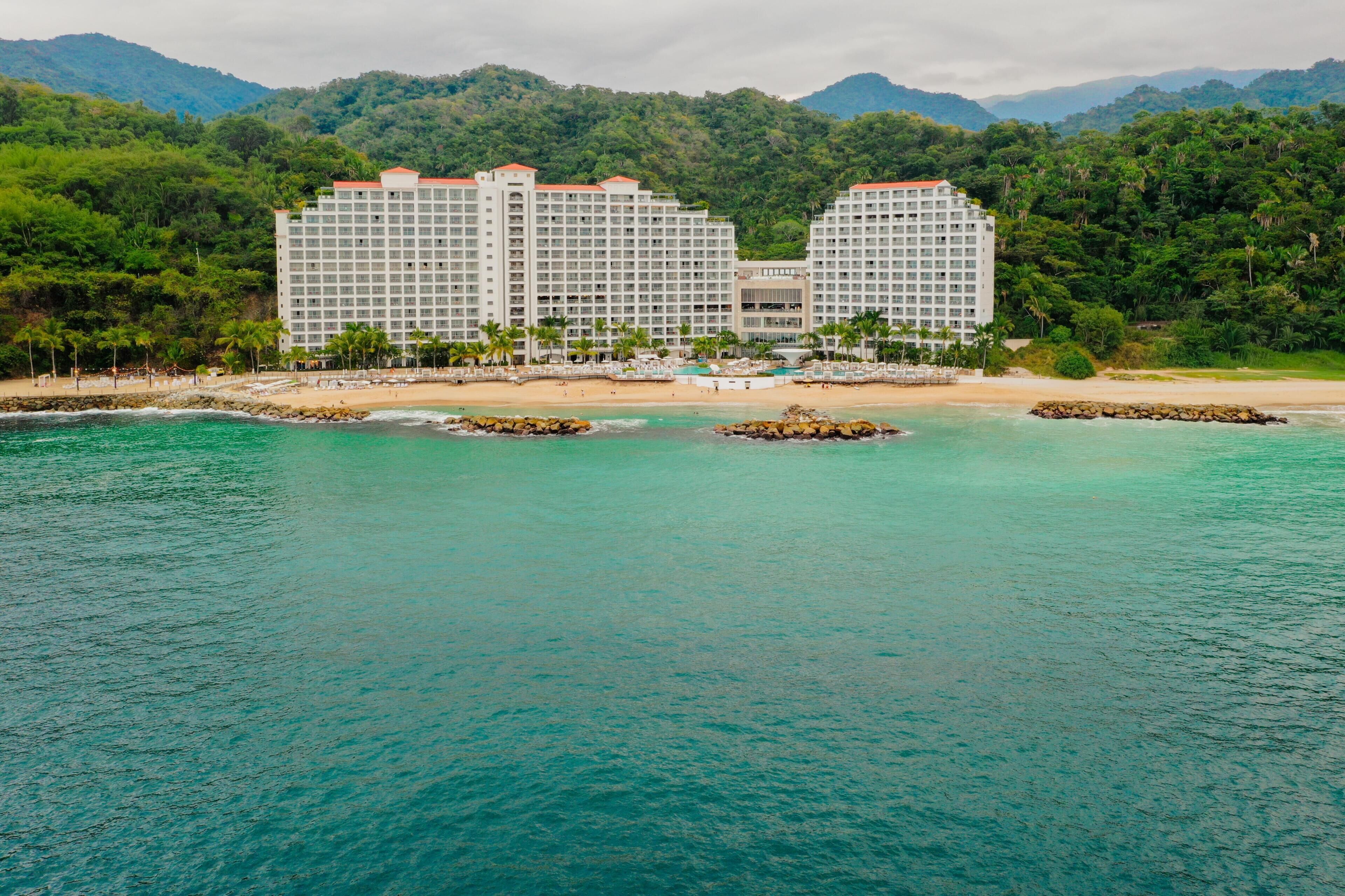Un hotel de varios pisos, con playa y montañas al fondo.