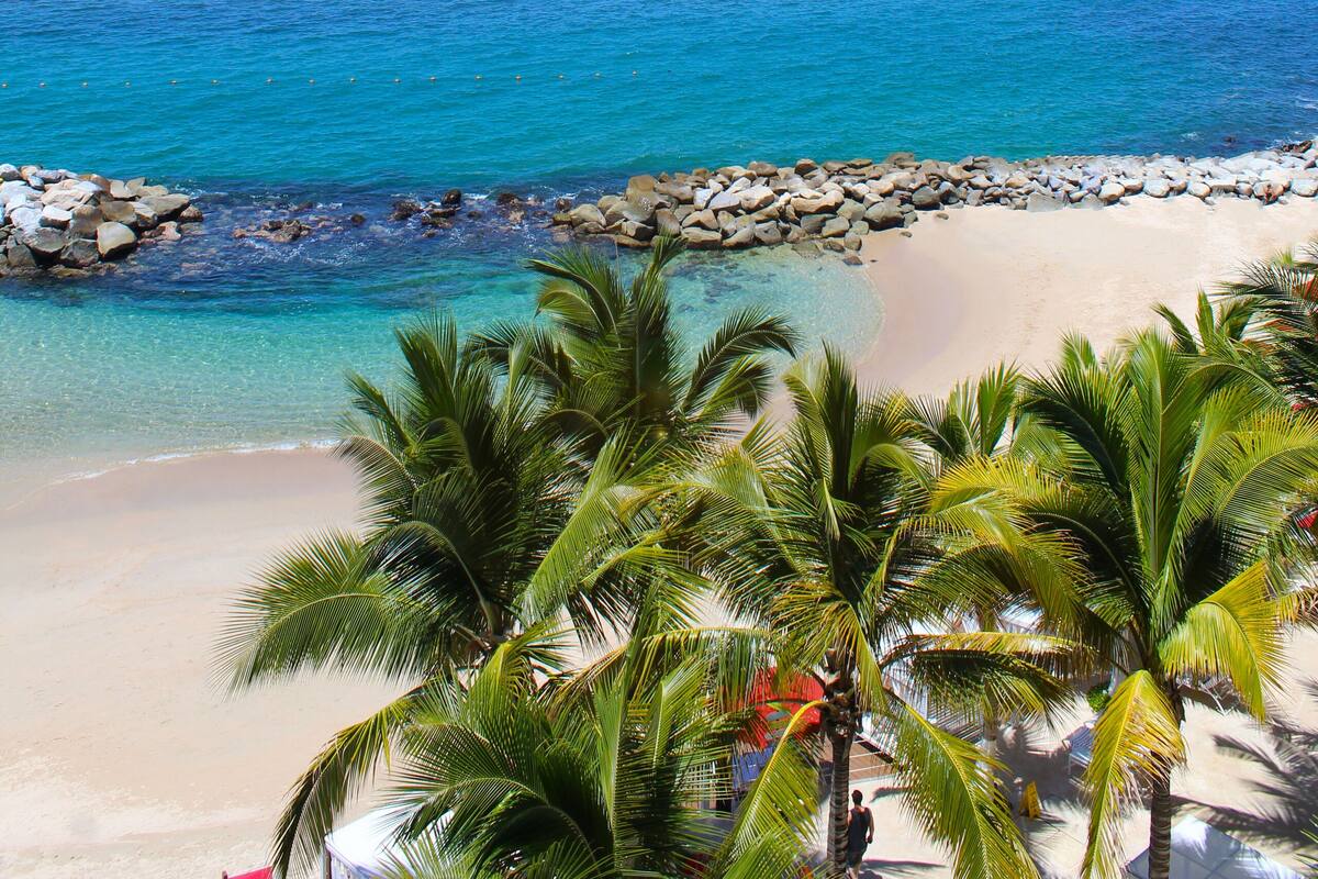 A beach with clear blue water, a rocky shoreline, and palm trees.