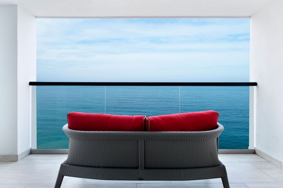 A balcony with a wicker chair and red cushions, overlooking a large body of water.
