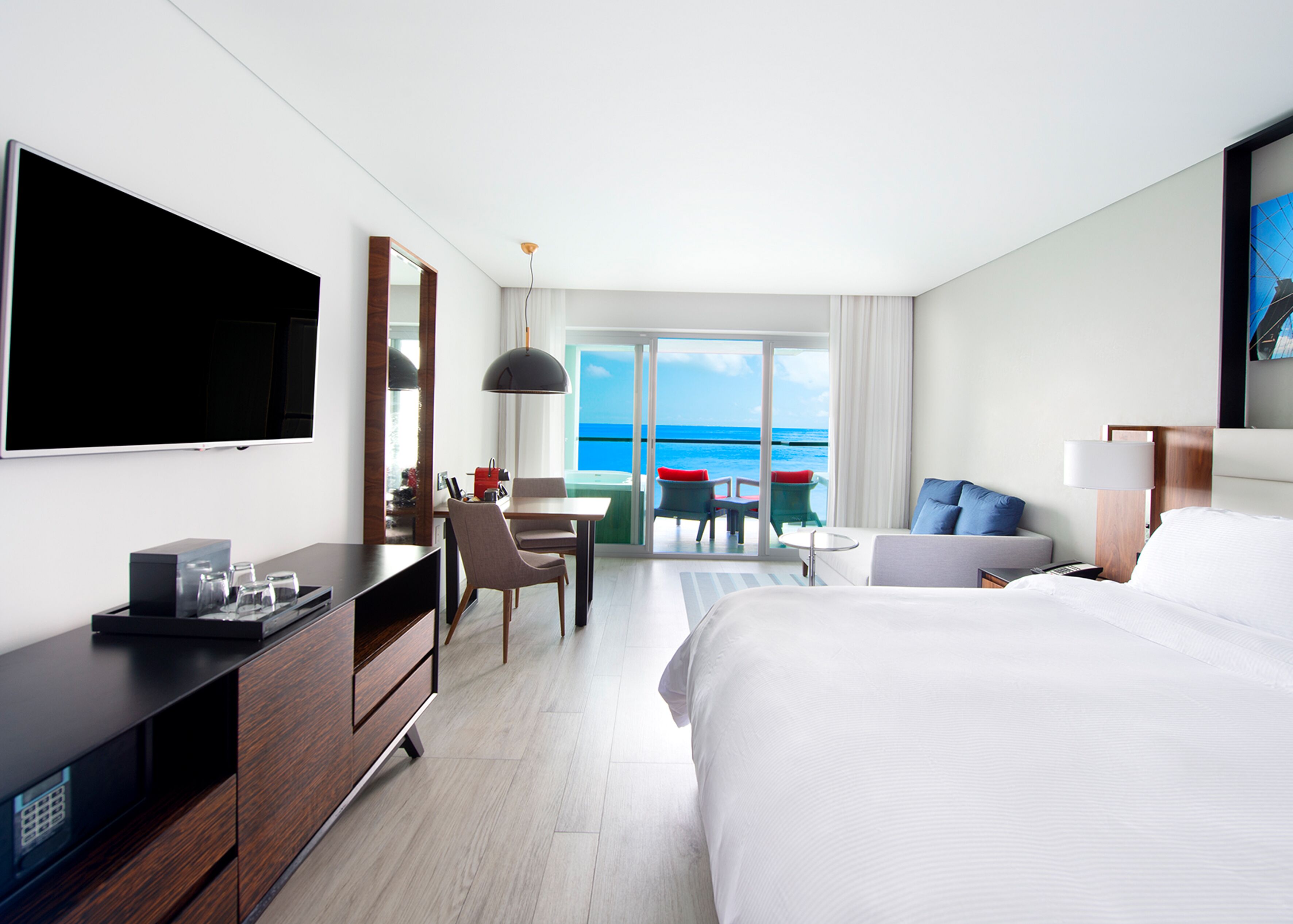 A modern hotel room with a large bed, a desk with a flat-screen TV, a dining area with chairs, and a balcony with a view of the ocean.