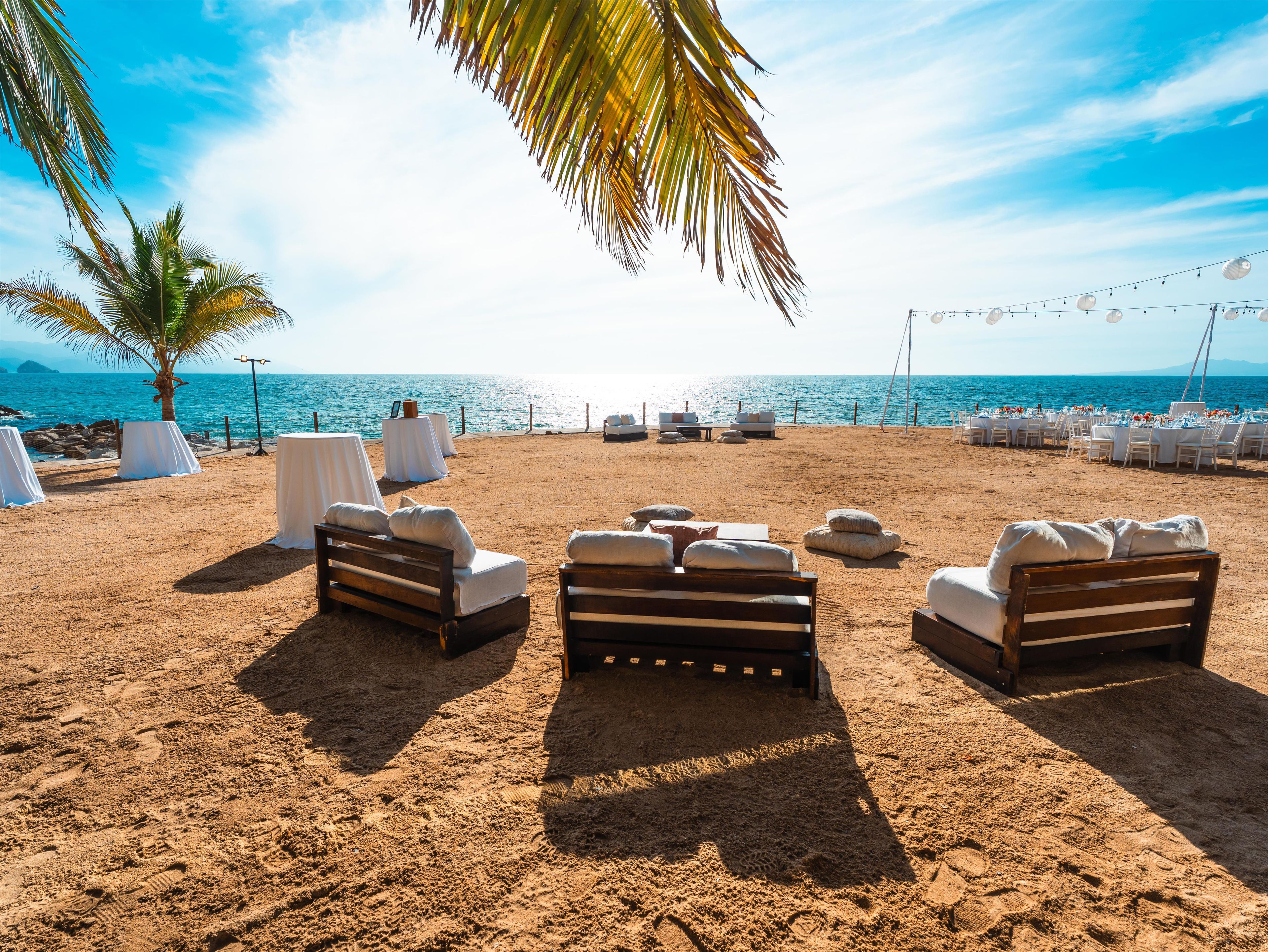 Decoración para una boda en la playa, con mesas blancas, sillas acolchadas y una palmera.