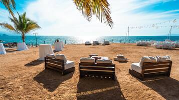 A beach wedding setup with white tables, cushioned chairs, and a palm tree.