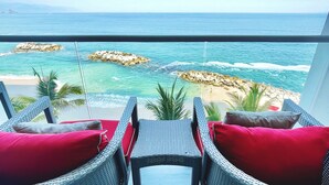 Un balcón con sillas de mimbre y cojines rojos, con vista a una playa con formaciones rocosas y agua azul cristalina.