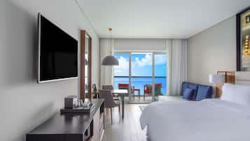 Habitación de hotel con cama, escritorio, televisor, zona de comedor con sillas y mesa, y balcón con vista al océano.