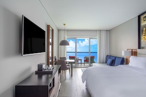 A hotel room with a bed, a desk, a TV, a dining area with chairs and a table, and a balcony with a view of the ocean.
