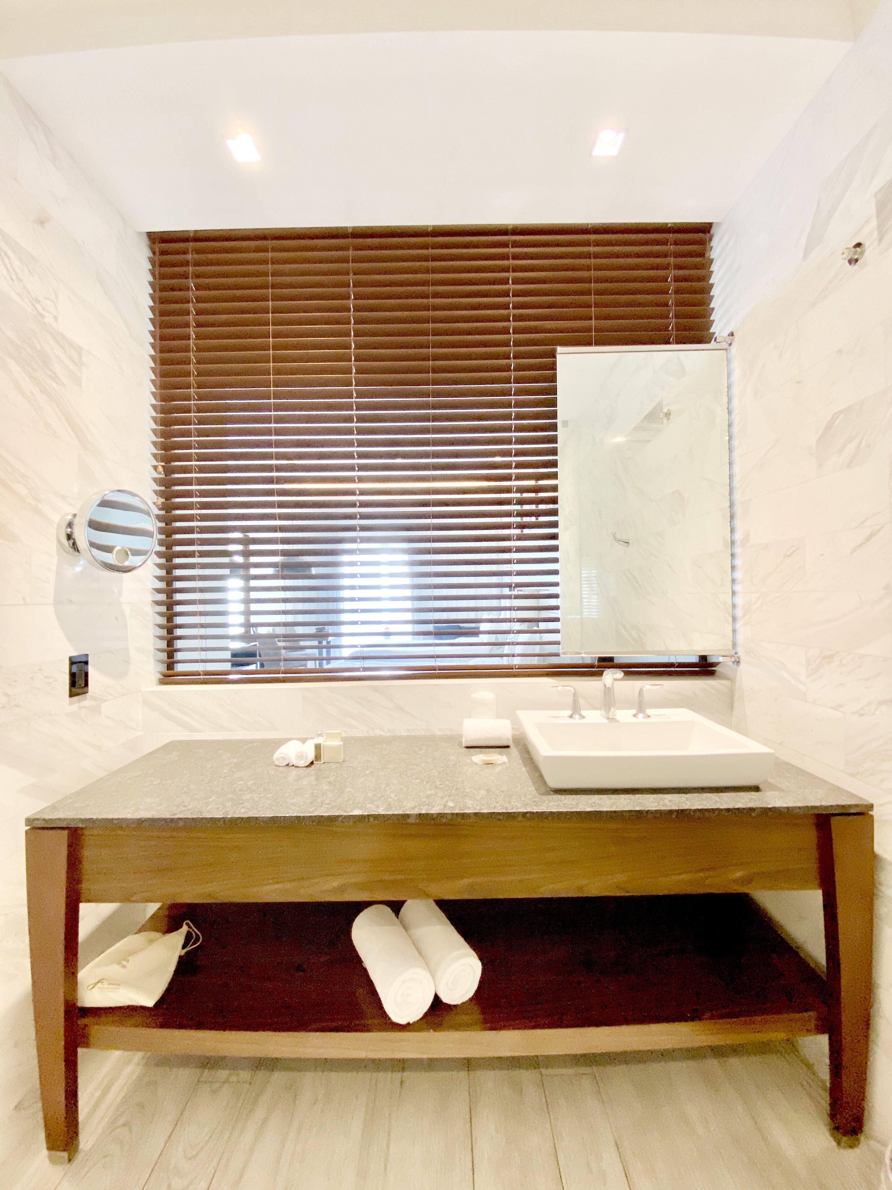 A modern bathroom with a wooden vanity, a large mirror, and a window with blinds.