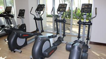 Une salle de sport équipée de divers vélos et tapis de course fixes, de grandes fenêtres et offrant une vue sur les palmiers à l’extérieur.