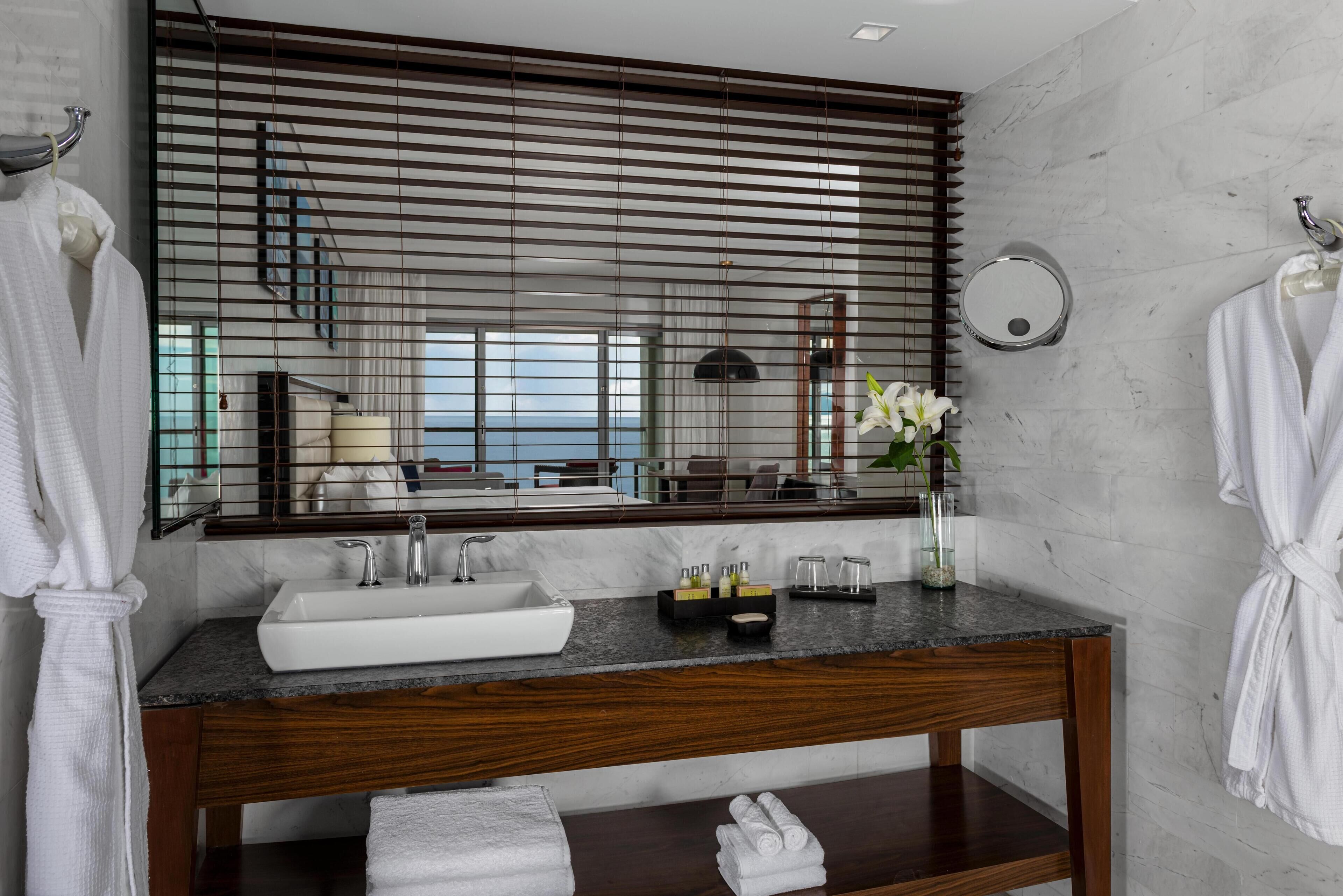A modern bathroom with a large mirror, wooden vanity, white sink, and a view of the sea through the window.