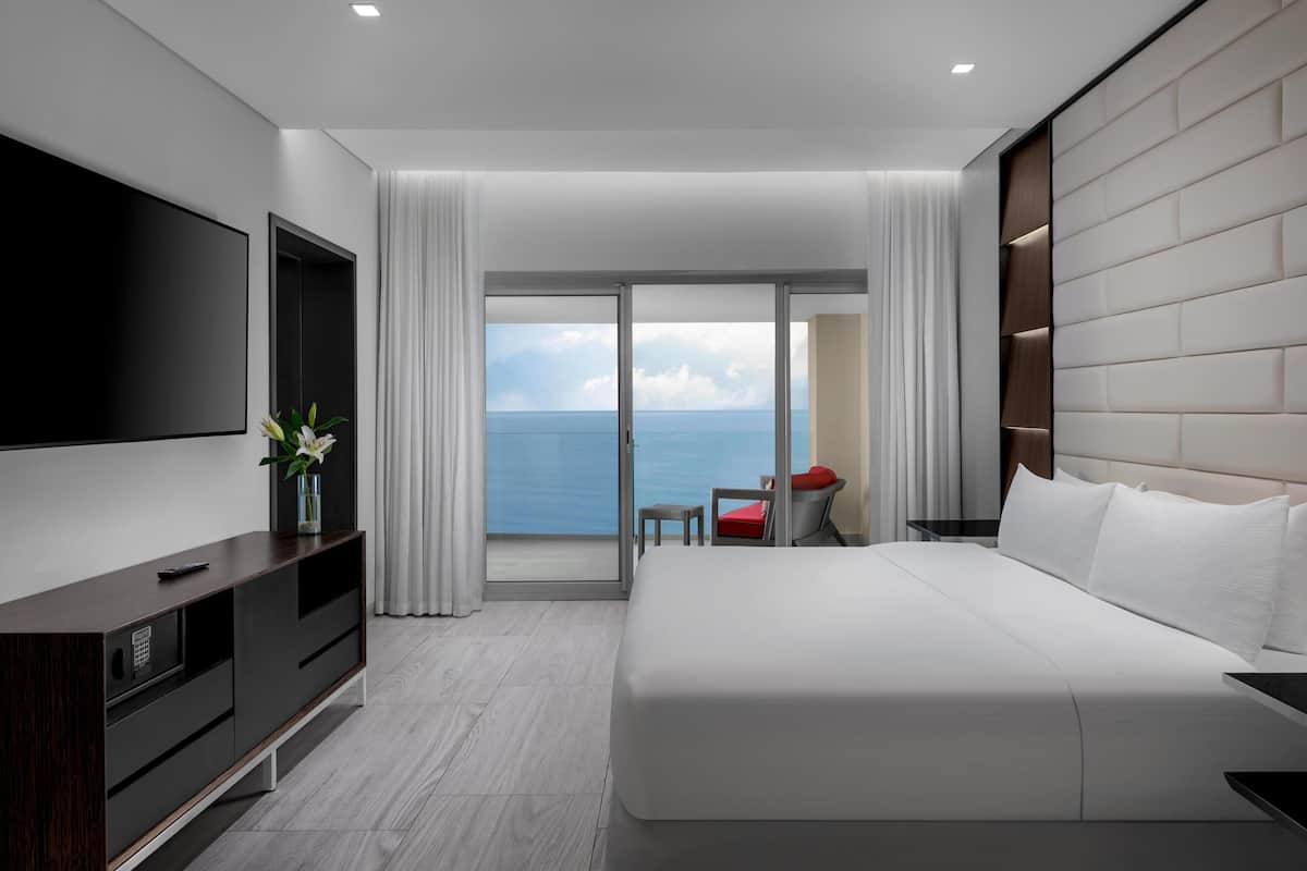 A modern hotel room with a large bed, a flat-screen TV, a small table with a red cushion, and a view of the sea through a glass door.
