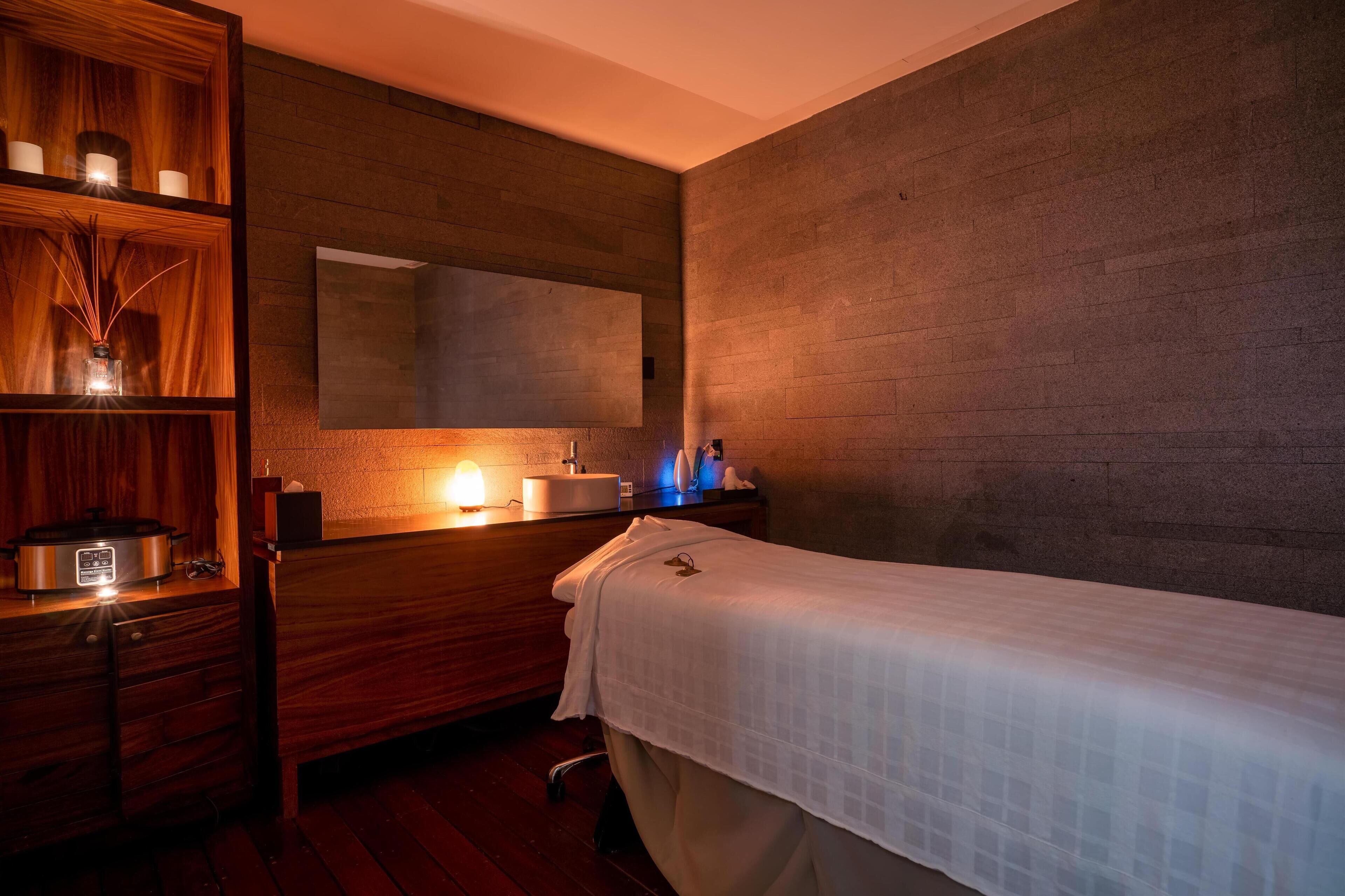 A spa room with a massage table, wooden shelves with candles, a mirror, and a modern sink.