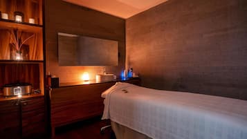 A spa room with a massage table, wooden shelves with candles, a mirror, and a modern sink.
