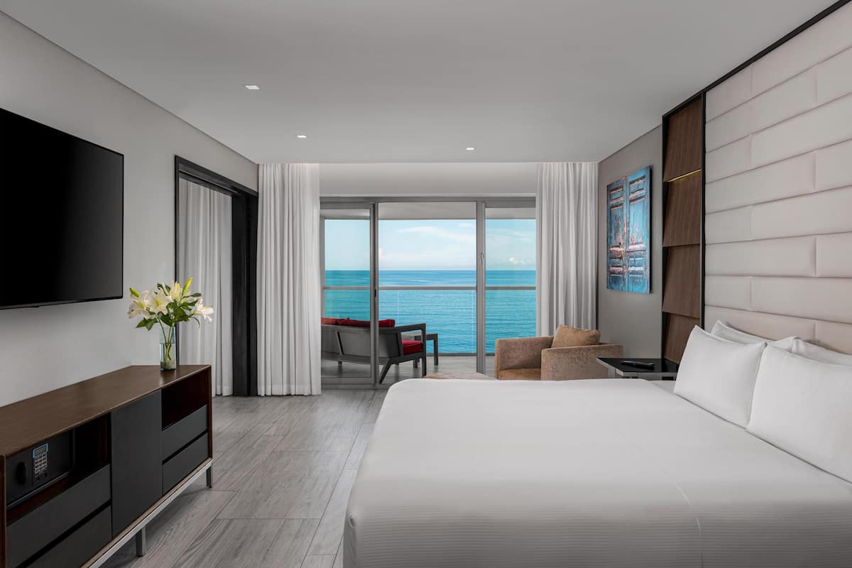 A modern hotel room with a large bed, a flat-screen TV, a wooden dresser, a seating area with a view of the ocean, and a large window.