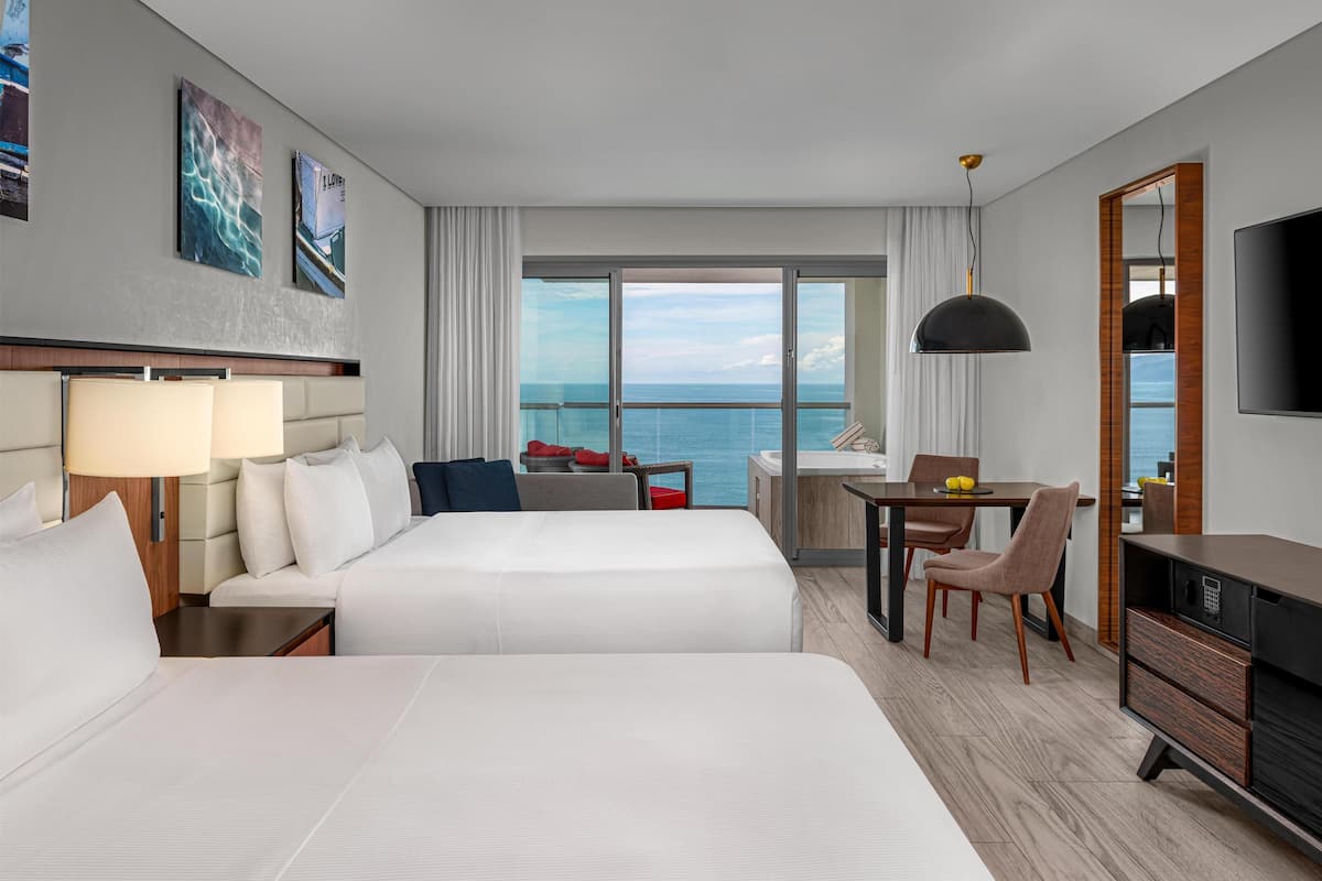 A hotel room with two beds, a dining area, a TV, and a view of the ocean.