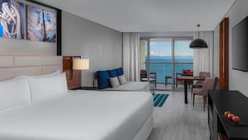 A modern hotel room with a large bed, a sofa, a dining area with a table and chairs, and a view of the sea.
