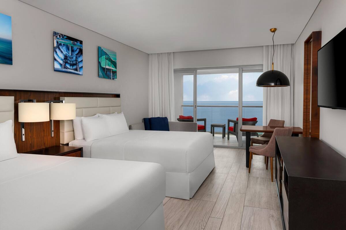 A hotel room with two beds, a desk, a TV, and a balcony with a view of the sea.