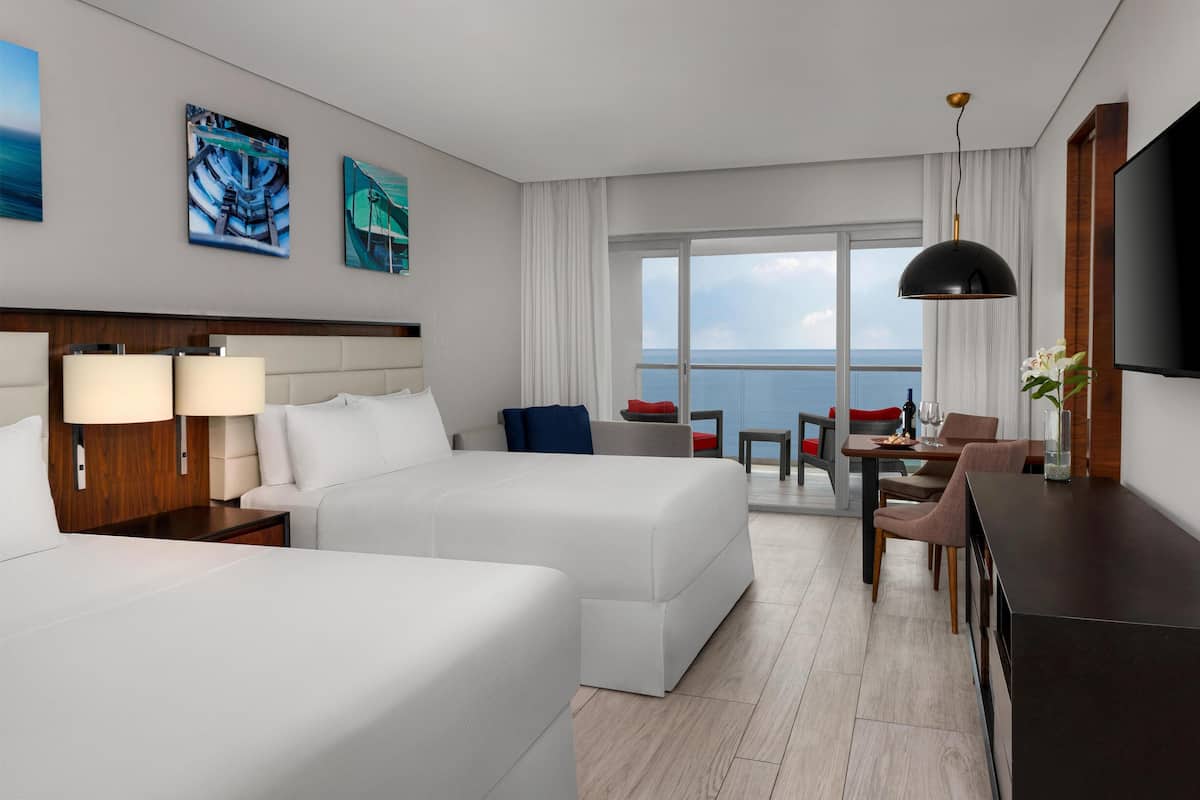 A hotel room with two beds, a desk, a chair, a TV, and a balcony with a view of the sea.