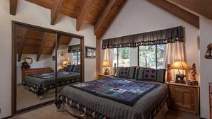 A bedroom with a large mirror, a bed with a quilt, a window with curtains, and a wooden ceiling. - Pinnacle View (Truckee)