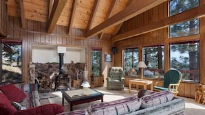 A rustic cabin interior with a stone fireplace, wooden furniture, and large windows offering views of the surrounding forest. - Pinnacle View (Truckee)