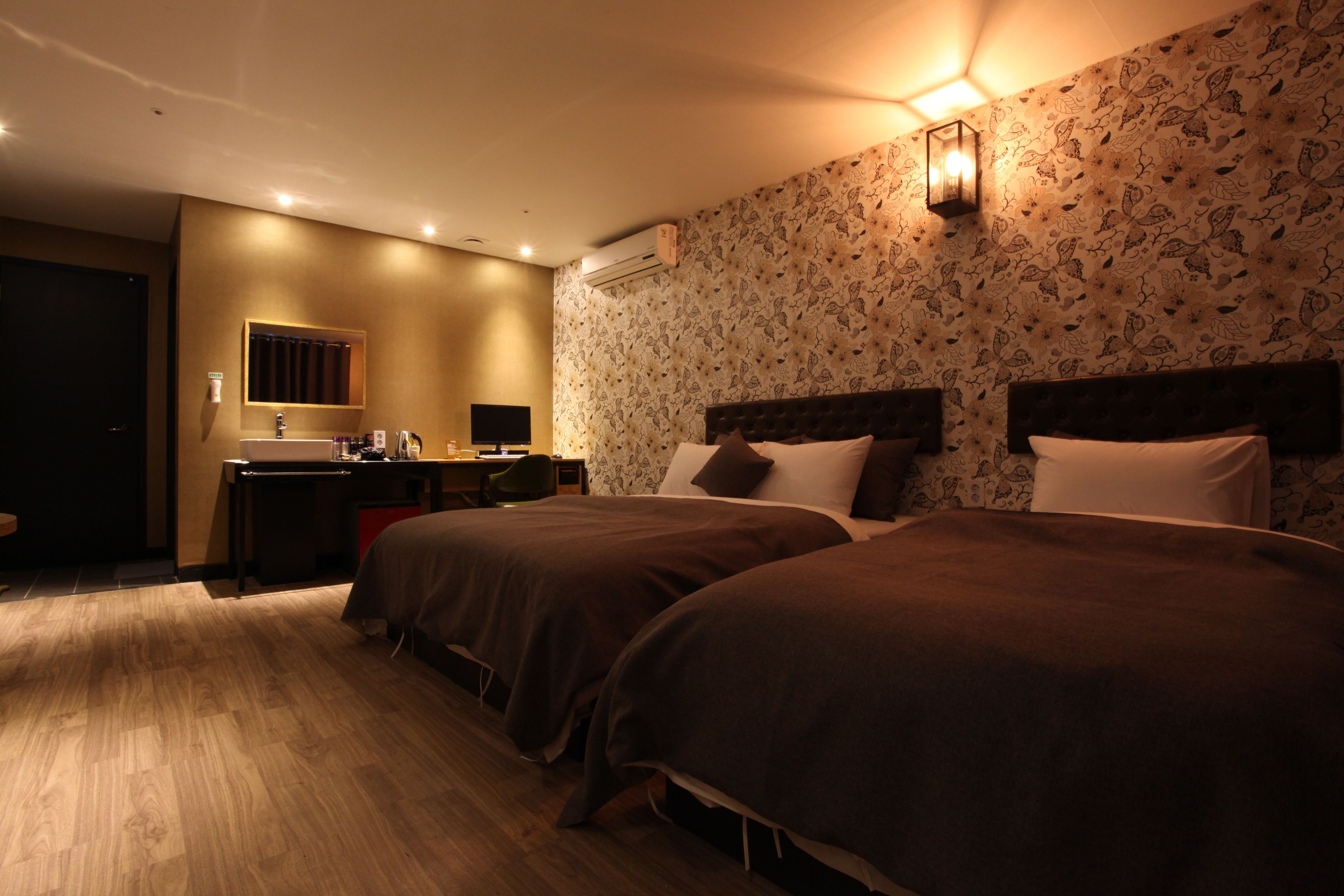 A hotel room with two beds, a desk, a chair, and a wall-mounted air conditioner.