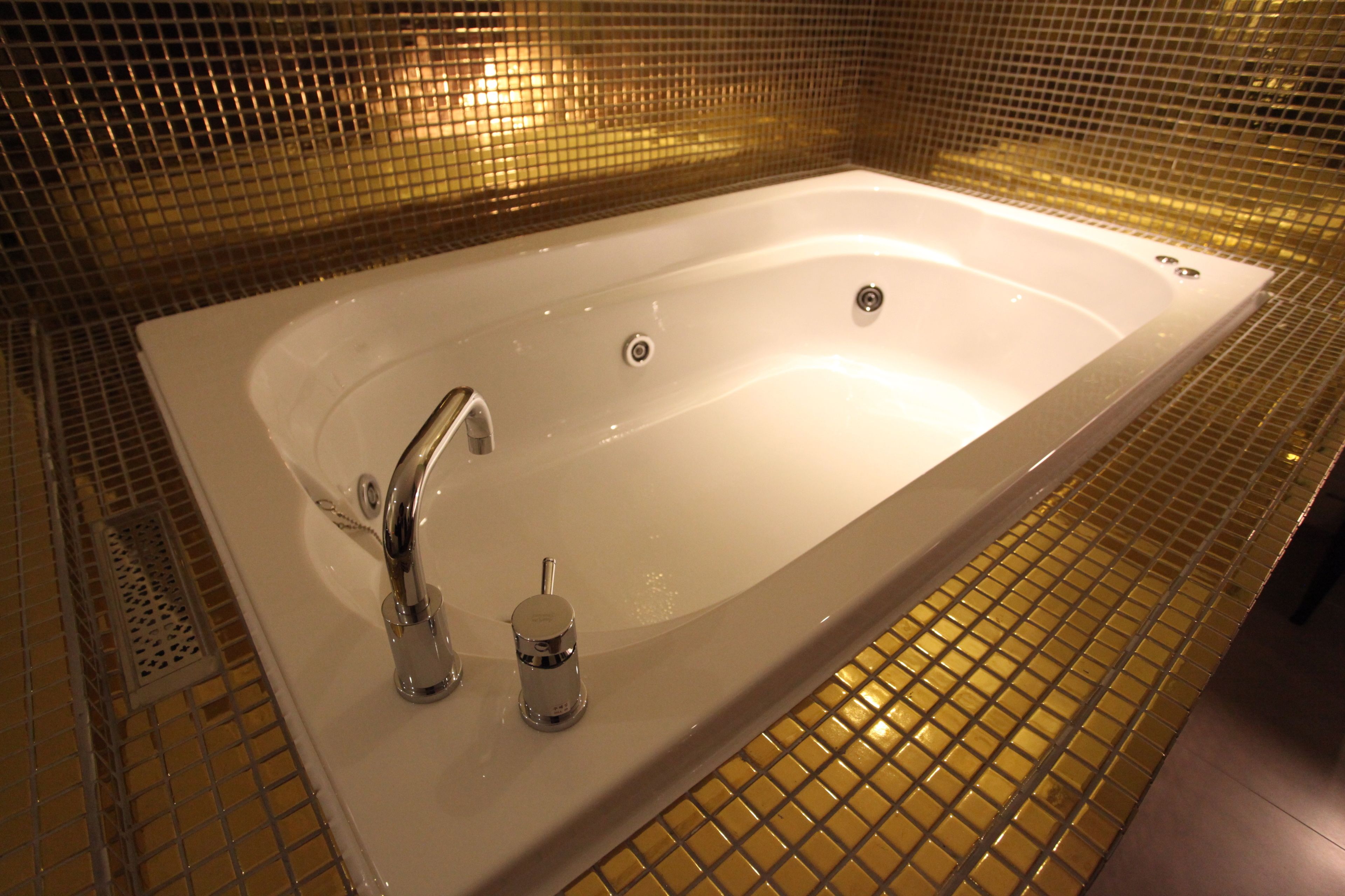 A rectangular bathtub with a single faucet and a handheld showerhead, surrounded by a tiled floor and walls.