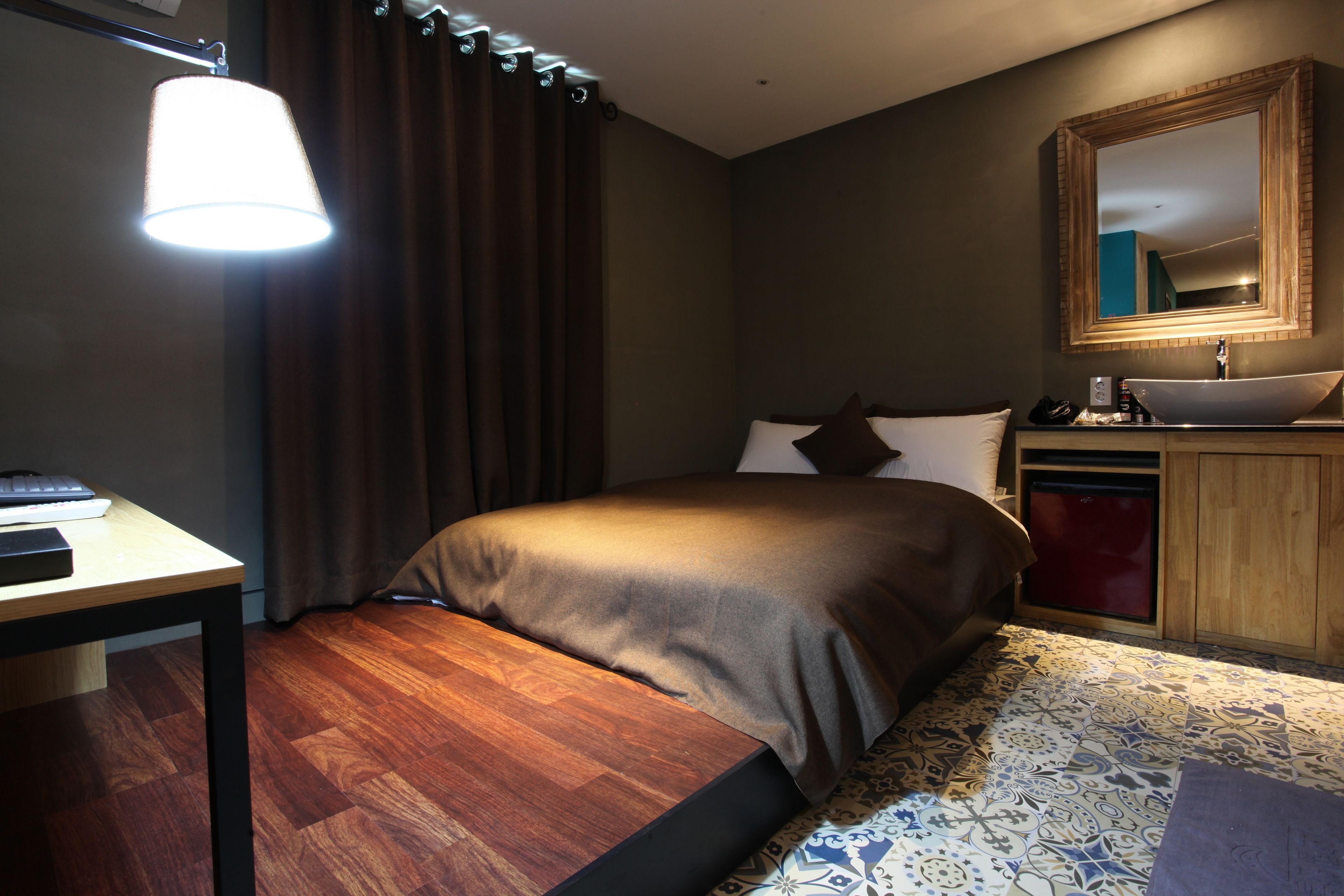 A hotel room with a bed, bedside table, lamp, mirror, and a small sink.
