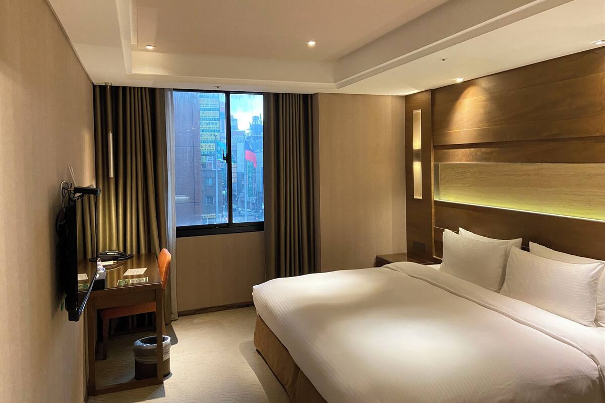 A hotel room with a large bed, a desk with a chair, a TV, and a window with curtains.