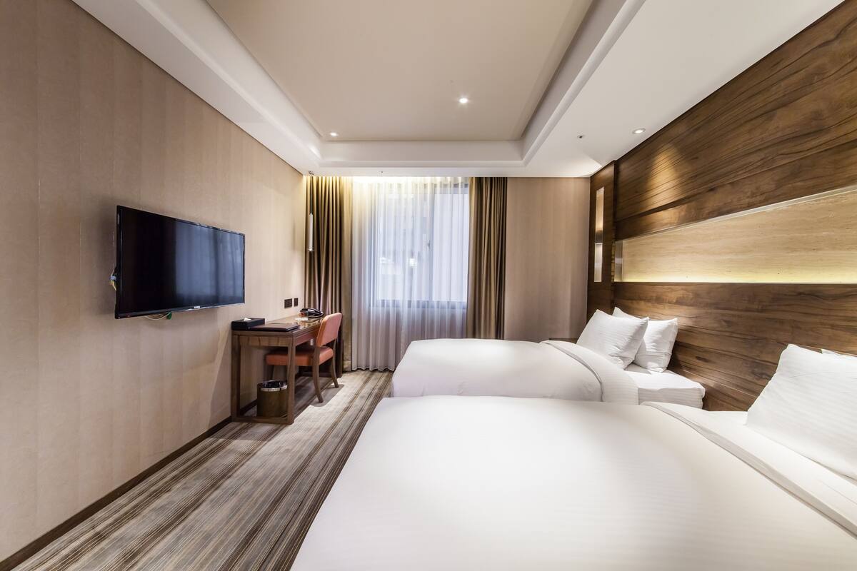 A hotel room with a flat-screen TV mounted on the wall, a desk with a chair, two beds with white linens, and a large window with curtains.