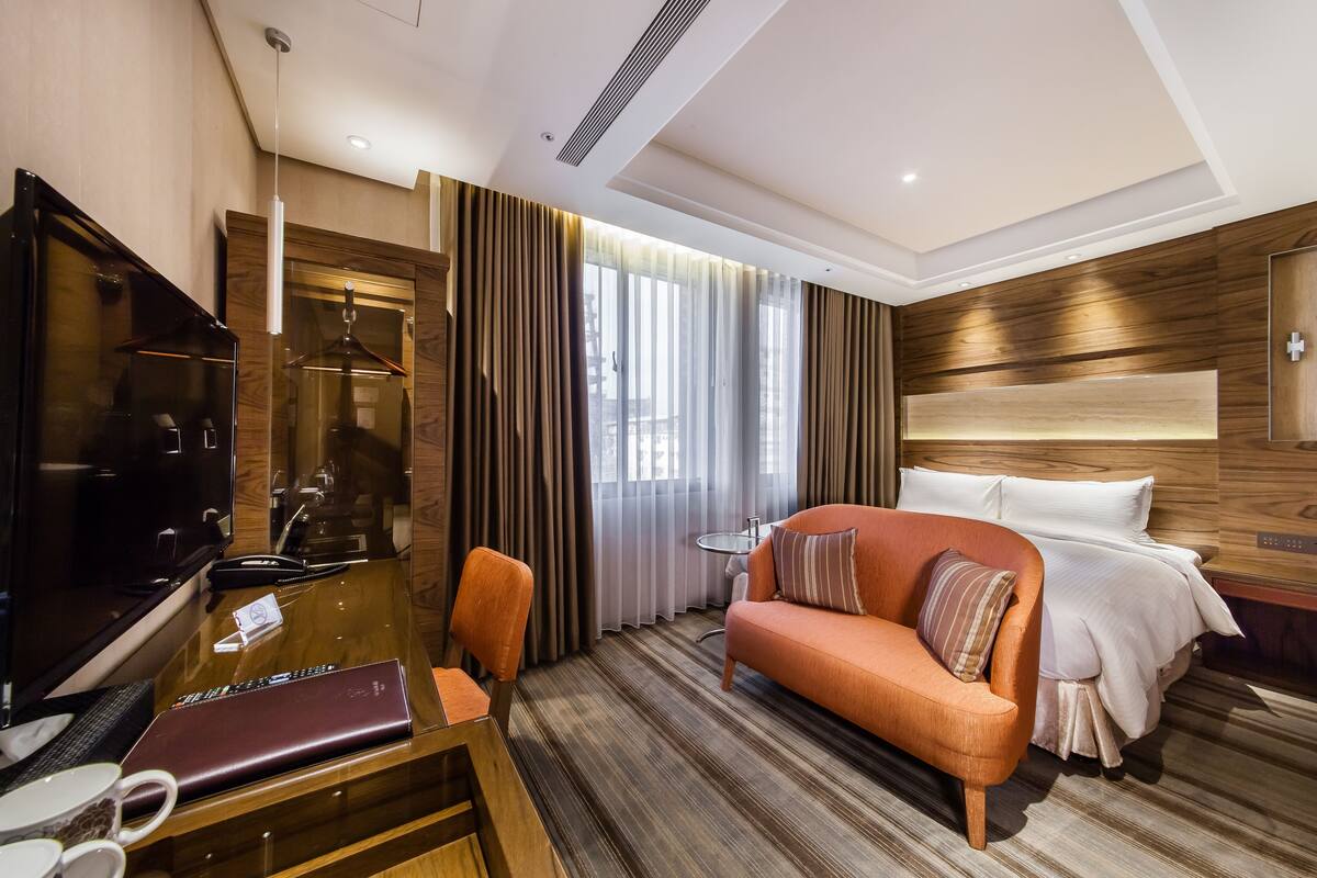 A hotel room with a wooden headboard, a sofa, a TV, a desk with a laptop, and a bed with white bedding.
