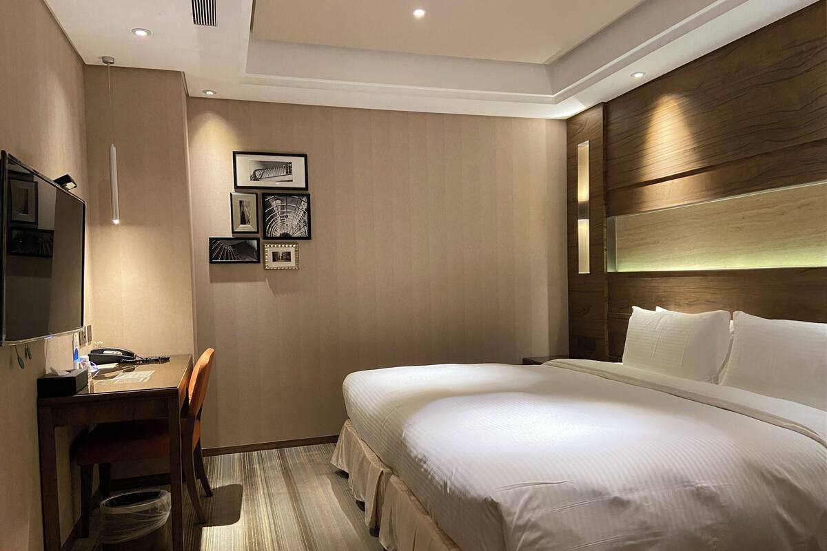 A hotel room with a large bed, a desk with a chair, a trash bin, and framed pictures on the wall.