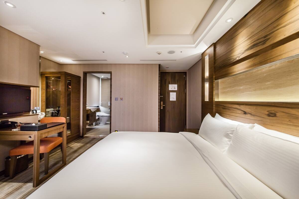 A hotel room with a bed, a desk with a chair, a television, and a wooden headboard.