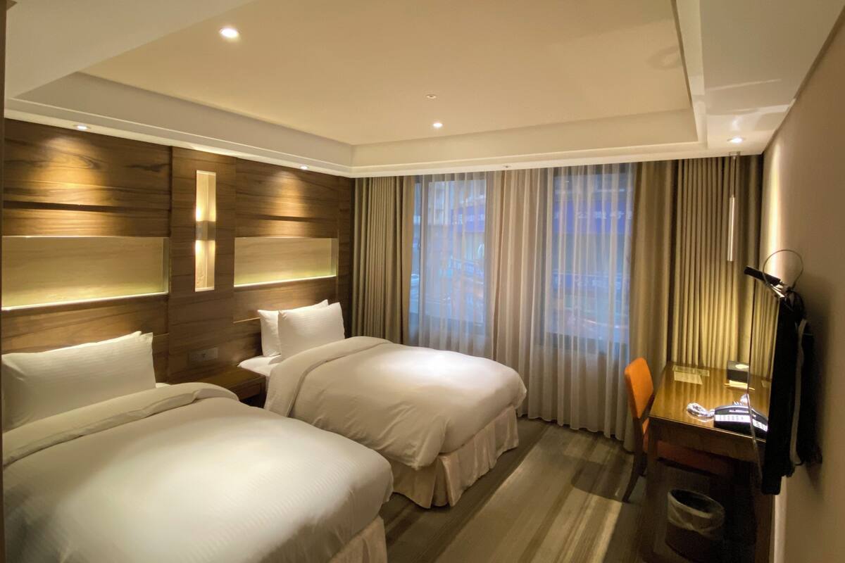 A hotel room with two beds, a desk, a chair, and a large window with curtains.
