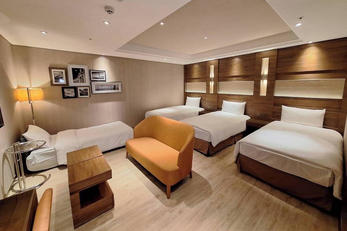 A hotel room with three beds, a sofa, a small wooden table, and a lamp.