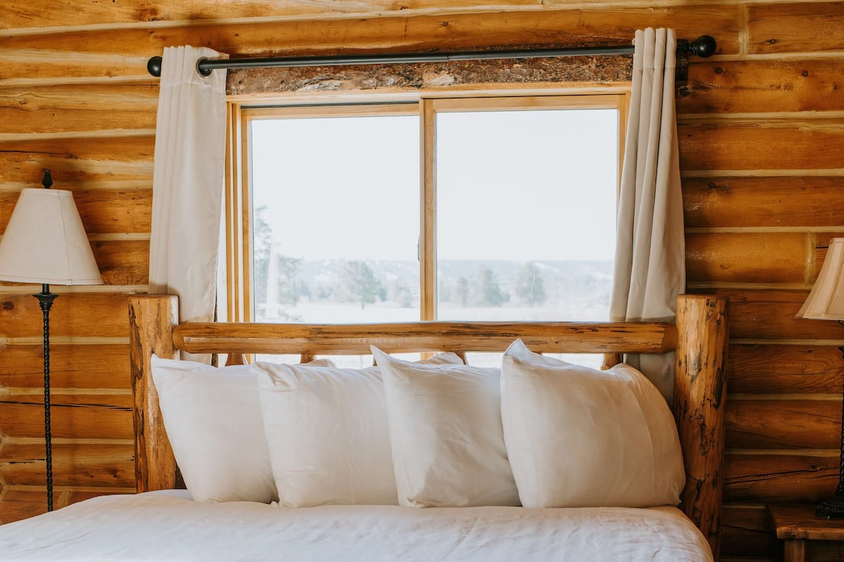 A wooden cabin-style room with a bed, pillows, a window, and a lamp.