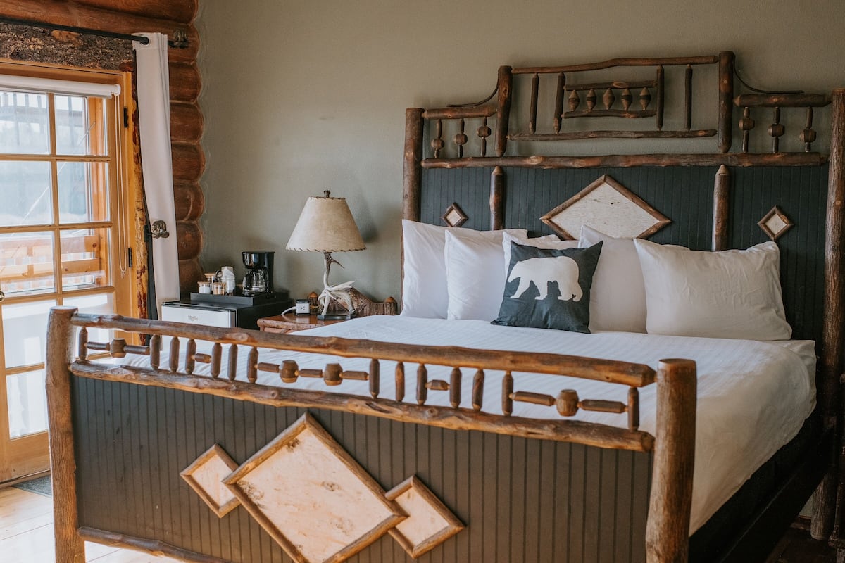 A wooden bed with a unique headboard design, a bedside table with a lamp, and a window with curtains.