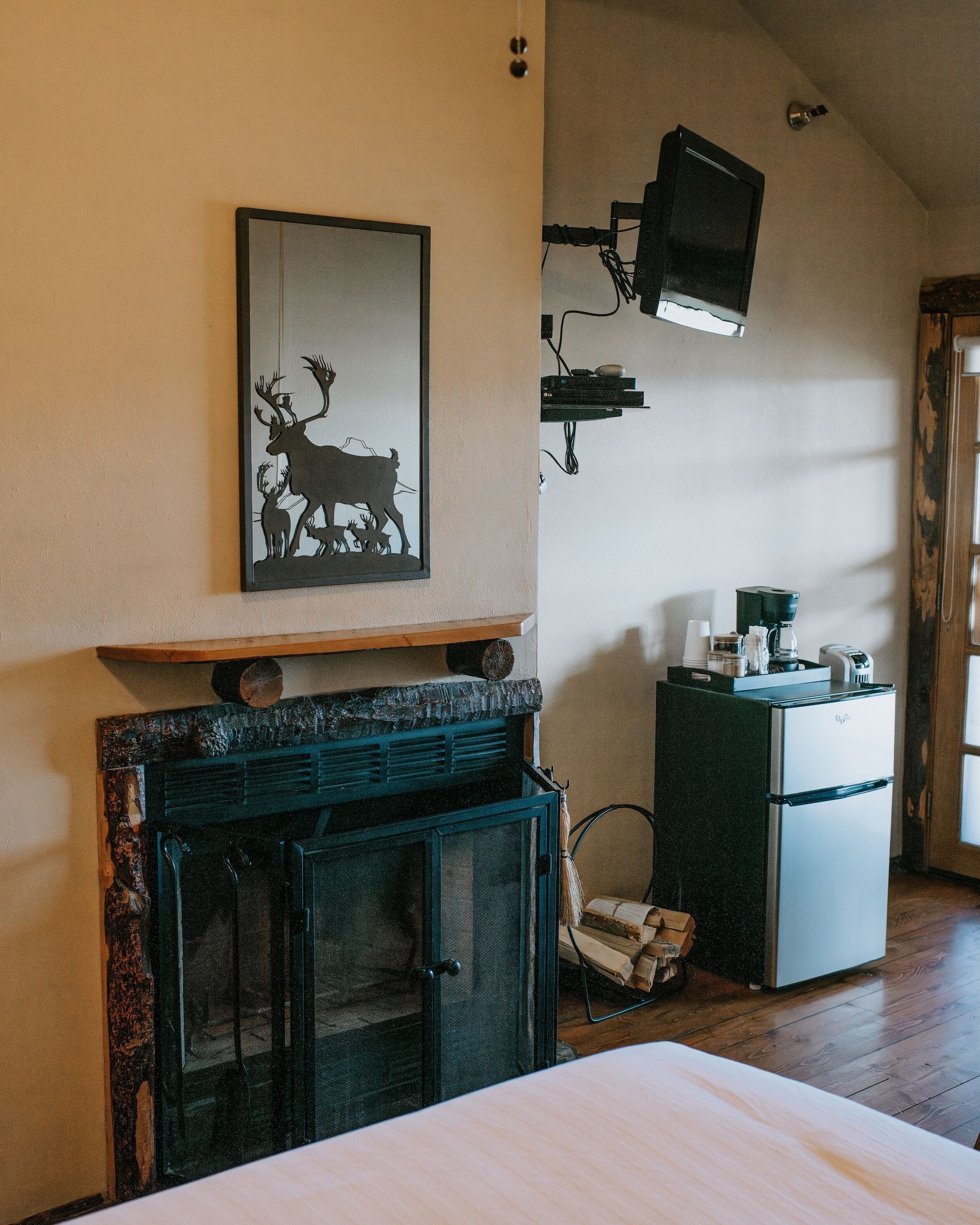 A room with a fireplace, a wall-mounted television, a refrigerator, and a framed picture of deer.