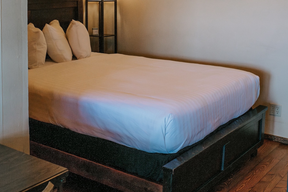 A bed with a wooden frame and a white mattress in a room with a nightstand, a lamp, and a framed picture on the wall.