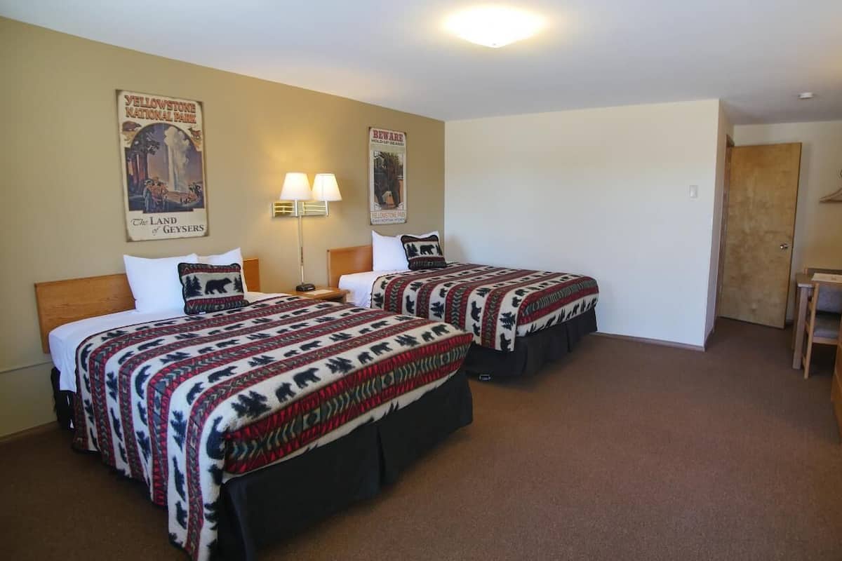 A room with two beds, each with patterned bedding, a wooden headboard, a nightstand with a lamp, and wall posters.