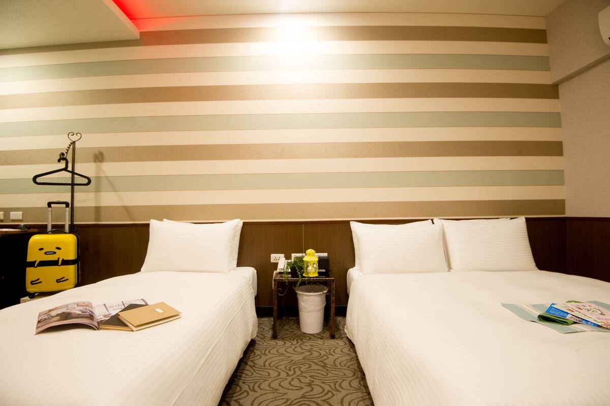 A hotel room with two beds, a small table with a trash bin, a yellow suitcase, and a hanging coat rack.
